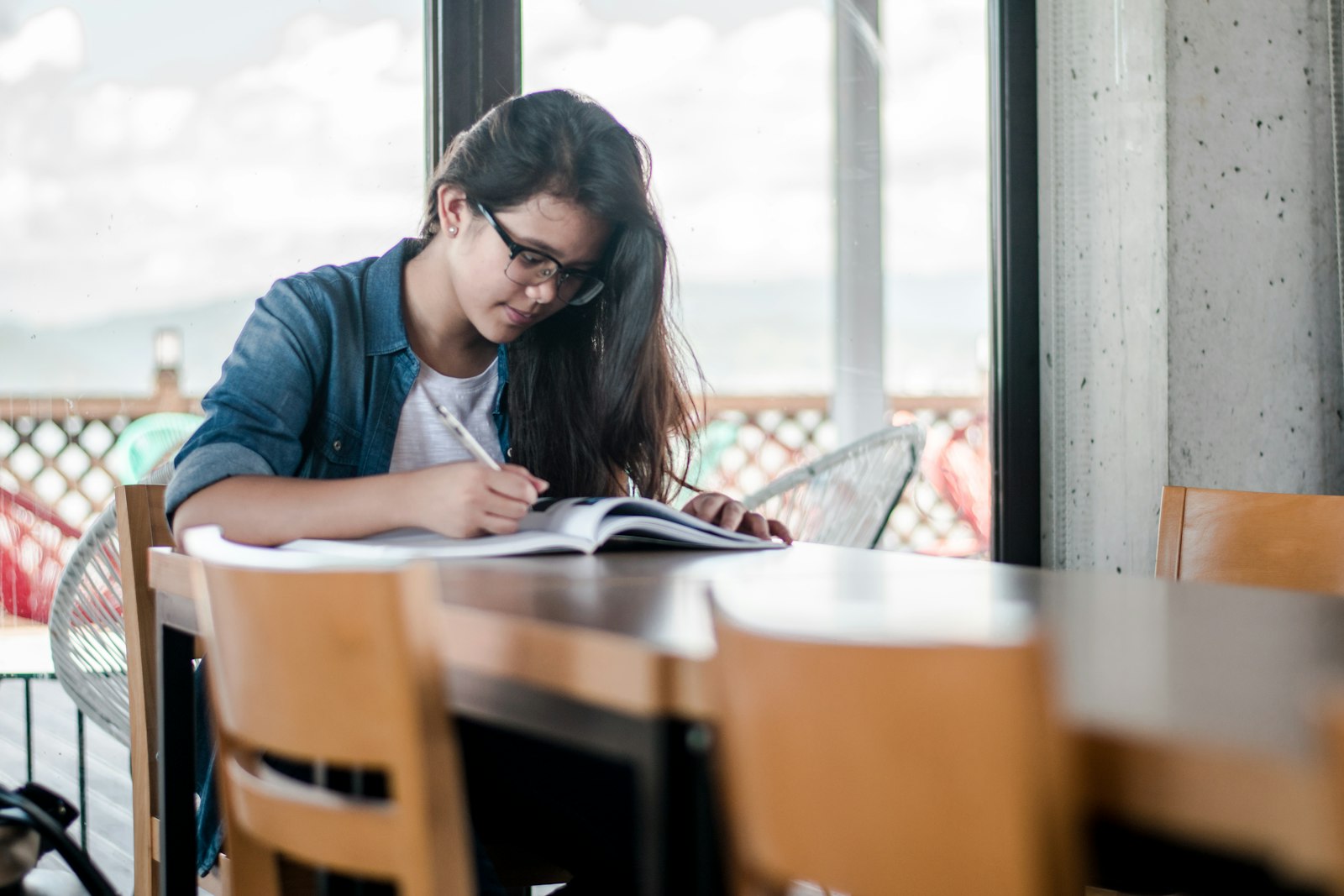 眼鏡をかけた少女が、窓辺のテーブルでノートに書き込んでいる